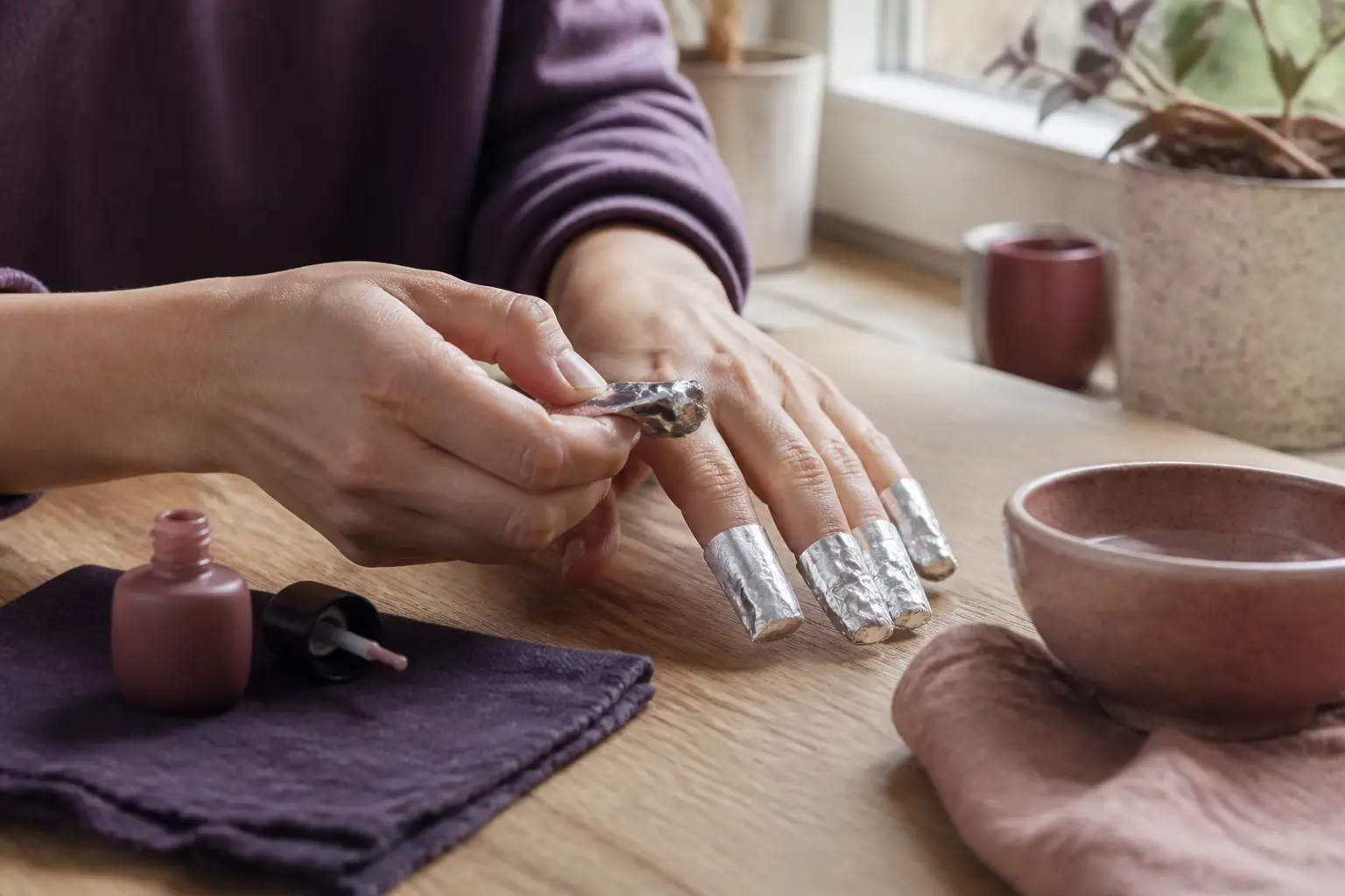 Mains retirant du vernis semi permanent avec papillotes en aluminium, sur une table de cuisine près d’une fenêtre, pour comment enlever du vernis semi permanent