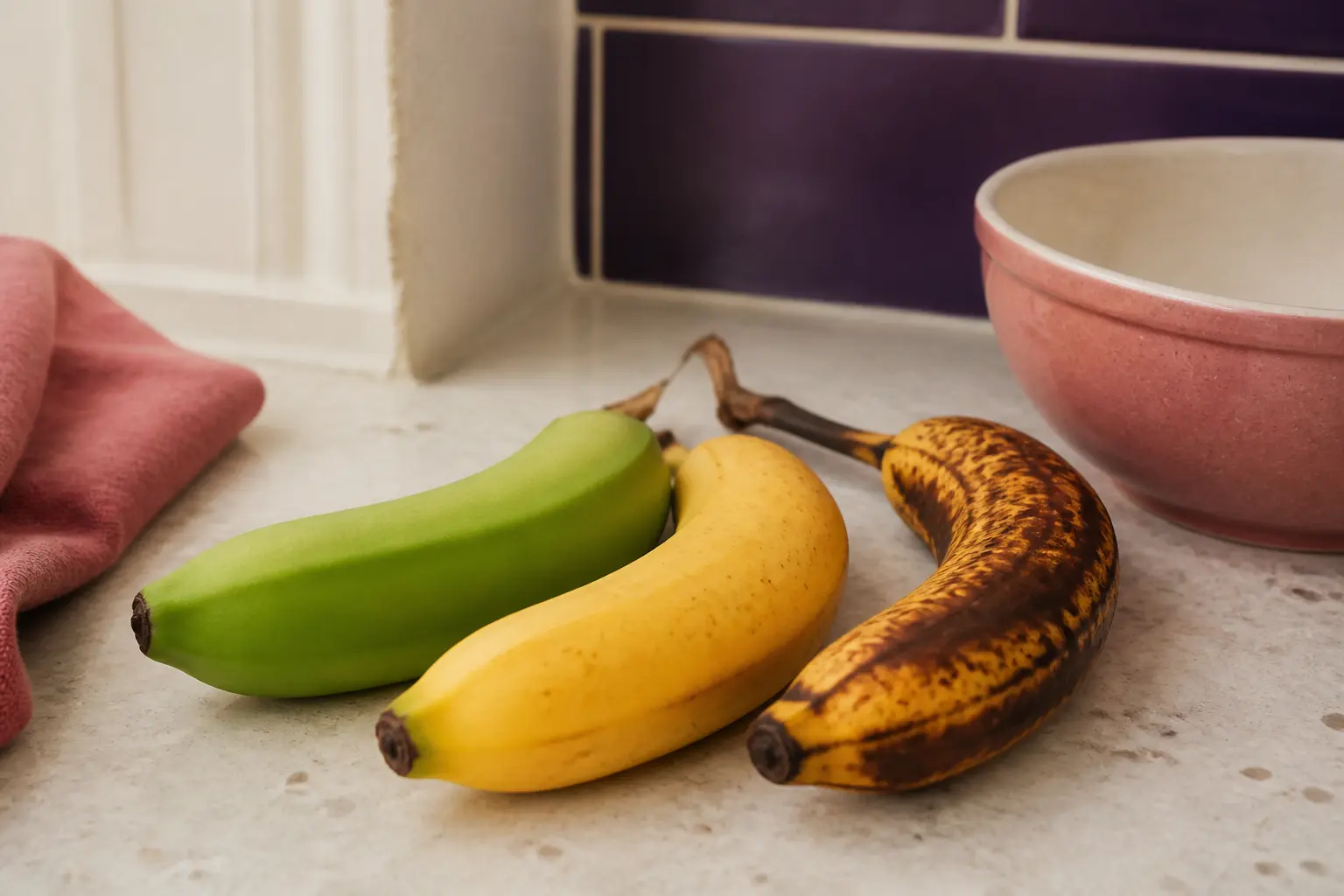 Banane constipation : trois bananes à maturité différente sur un plan de cuisine, de verte à très mûre, avec bol et torchon.